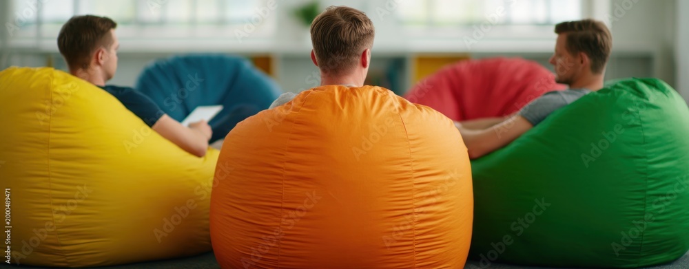 Fototapeta premium Diverse young men relaxing on colorful bean bag chairs in modern creative office during casual meeting reflecting informal collaboration and startup culture environment