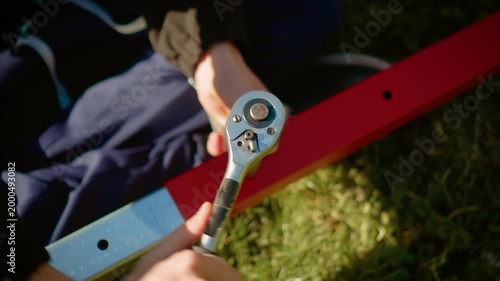 Manual worker tightening bolt with ratchet wrench on red metal beam in outdoor setting. DIY construction assembly repair project with mechanic tool equipment and craftsmanship detail in sunlight. Hand