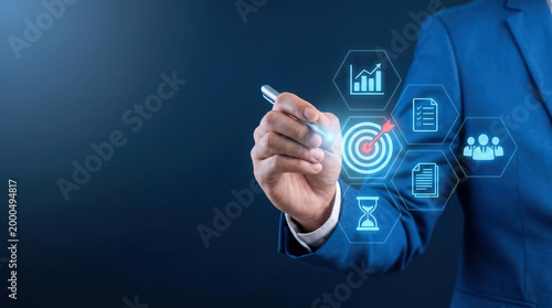 A man in a suit pointing with a pen at a digital display showing target, graphs, hourglass, and team icons.