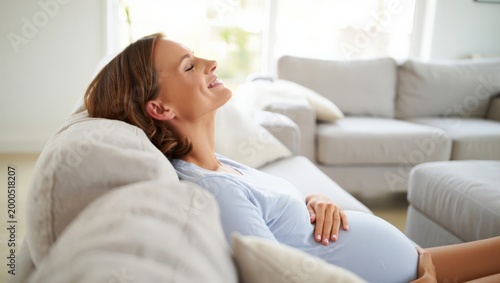 Free Download A pregnant woman experiencing stomach pain lies on a couch in a bright living room, clutching her stomach with a look of discomfort on her face. - GFXLoad