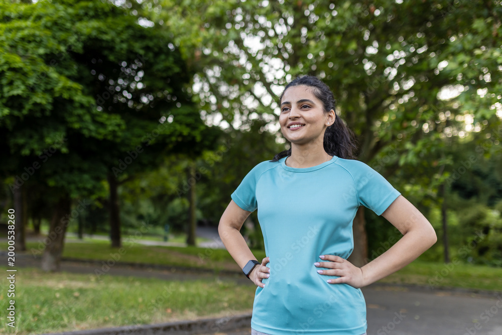 Fototapeta premium Young indian woman in sportswear stands with hands on hips, smiling and looking away in a green urban park, enjoying a confident, healthy, active lifestyle and fresh air