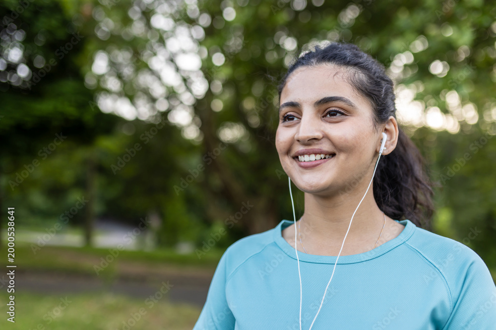 Naklejka premium Young indian woman smiling cheerfully outdoors, listening to music with earphones, enjoying fitness and a healthy lifestyle in a green park environment