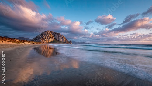 A serene coastal landscape features Morro Rock reflected in the wet sand at sunset.