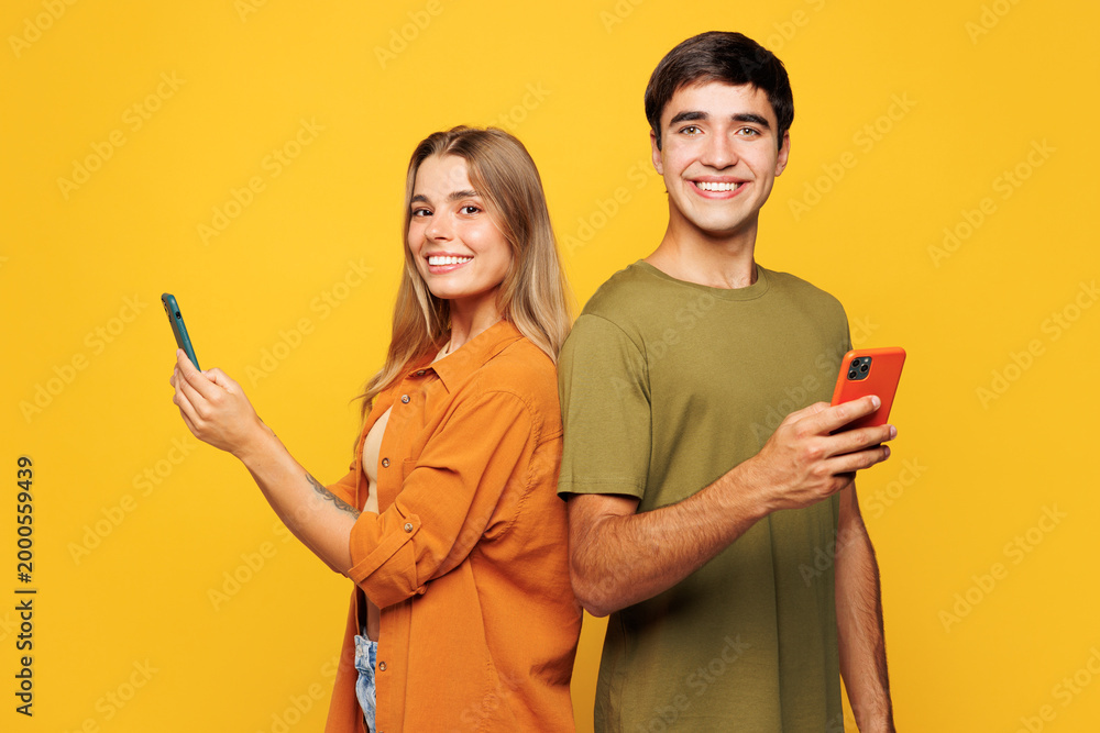 Obraz premium Side view smiling young couple two friends family man woman wear casual clothes hold in hand use mobile cell phone together stand back to back isolated on pastel plain light yellow background studio.