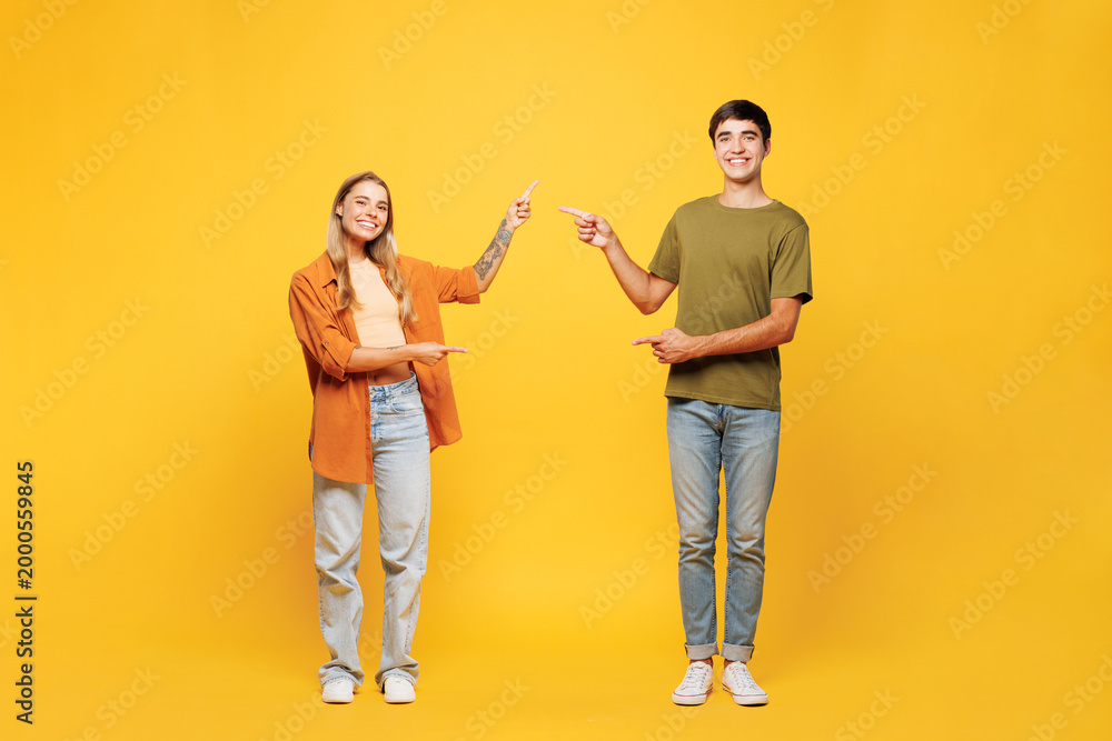 Fototapeta premium Full body fun young couple two friends family man woman wear casual clothes together point index finger between them on workspace area mock up isolated on pastel plain yellow color background studio.
