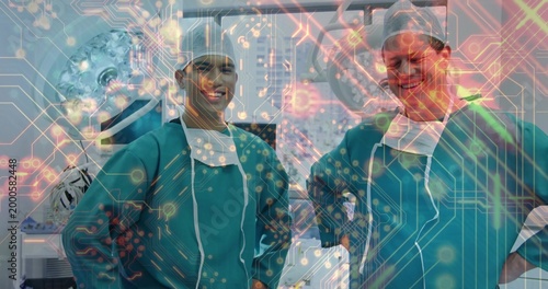 Smiling two surgeons in teal gowns, caps posing in OR, masks-lowered, screens, tech-overlay showing