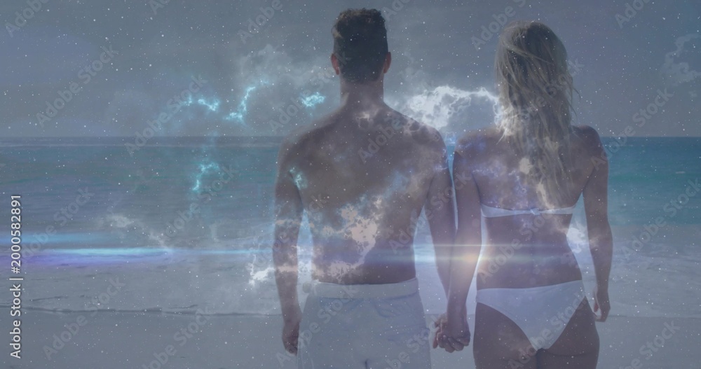 Fototapeta premium Holding hands couple facing ocean on shore in white swimwear, starry nebula and lens flare