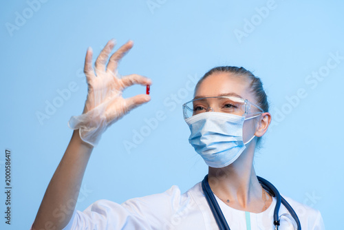 Medical professional wearing mask and safety glasses holding a red capsule, representing health, medicine, and research developing treatment