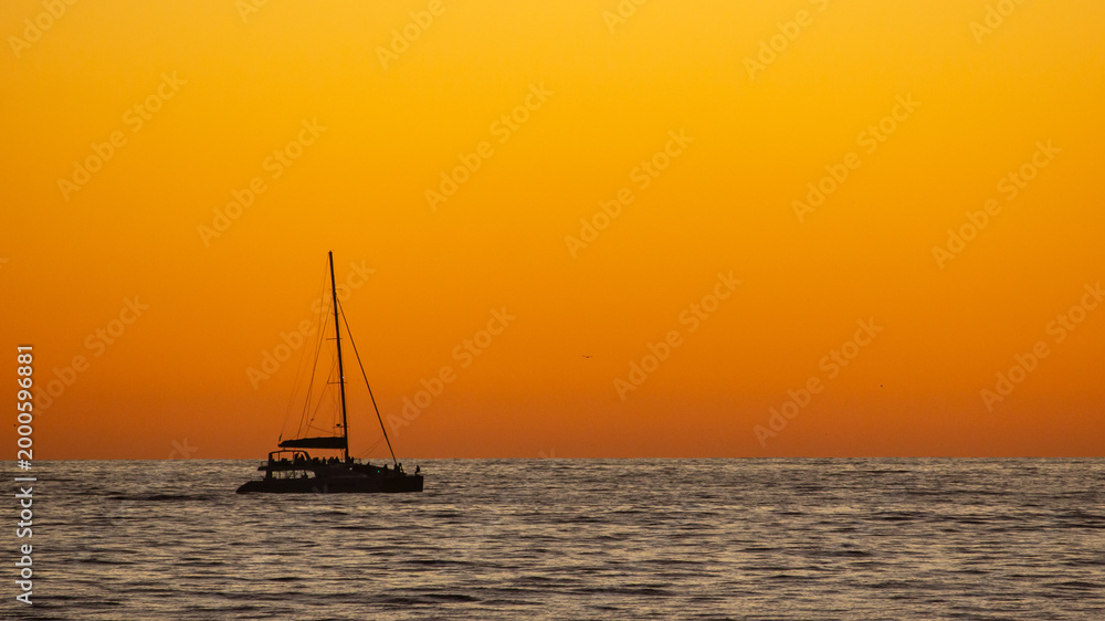 Fototapeta premium Silhouette of a distant Catamaran on the ocean against a vibrant golden orange sunset off the coast at Cape Town South Africa.