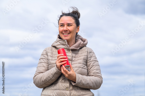 Smiling woman holding beverage can against cloudy sky.