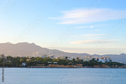 Coastal town with modern buildings nestled against rolling hills under a clear sky