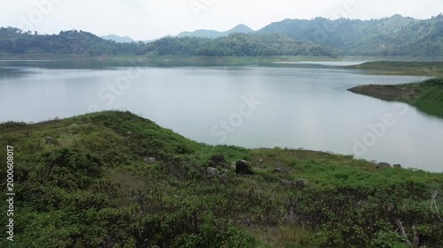 Aerial view flying over a serene reservoir lake surrounded by green grassy hills and distant mountains.