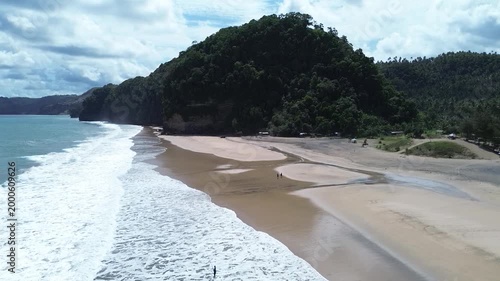 Cinematic drone view of a pristine seaside landscape, featuring gentle waves breaking on the shore and a massive forested headland.