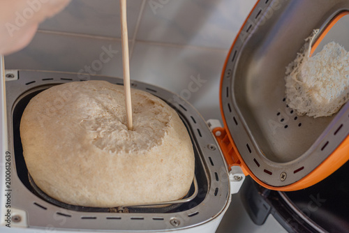 Unrecognizable person checking bread in electric breadmaking machine, closeup photo. Checking dough texture in bread machine with wooden stick, dough has risen too much