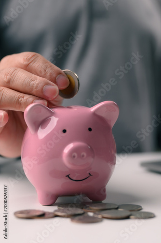 Adult hand inserts silver coin inside slot pink ceramic piggy bank sitting white table surrounded scattered change indoor setting representing financial planning retirement security investment.