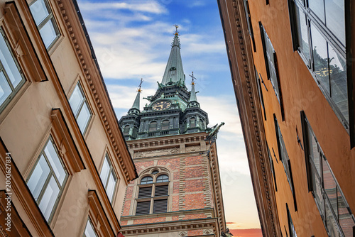 Tall green copper spire and brick clock tower of the German Church, framed by the narrow historic buildings of the Gamla Stan district in Stockholm, Sweden.
