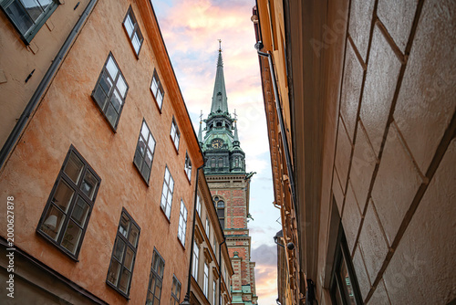 Tall green copper spire and brick clock tower of the German Church, framed by the narrow historic buildings of the Gamla Stan district in Stockholm, Sweden.