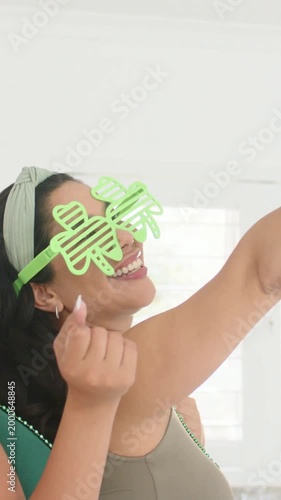 Vertical video: Smiling woman in green tank shamrock specs taking selfie in kitchen as cell appears