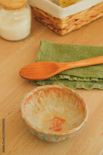 Kitchen scene with a wooden spoon resting on a green napkin, a ceramic bowl nearby, and a jar and woven basket on a wooden table, capturing home cooking moments