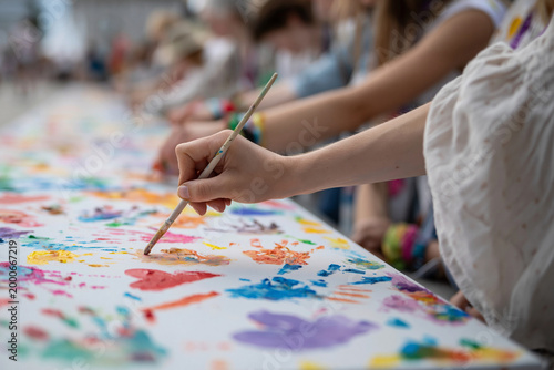 Hands paint vibrant colors on a large shared canvas. Fingers move confidently, blending hues with gentle strokes. Joyful creativity flows as colors explode across the surface