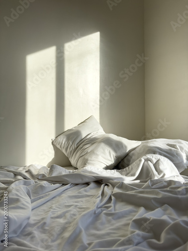Soft natural sunlight casting geometric window shadows on unmade white bed with pillows and wrinkled linen sheets in minimalist calm bedroom interior