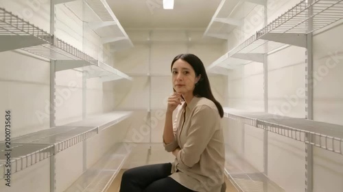 Sitting Asian woman in beige top, shifting gaze as camera rolls in pantry with shelves, pondering