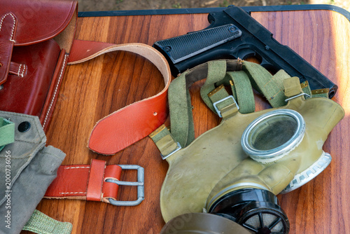 Set of vintage military equipment lying on a wooden table: gas mask, leather holster, belt, and pistol. Collectible historical items in retro style, daylight illumination, close-up shot.