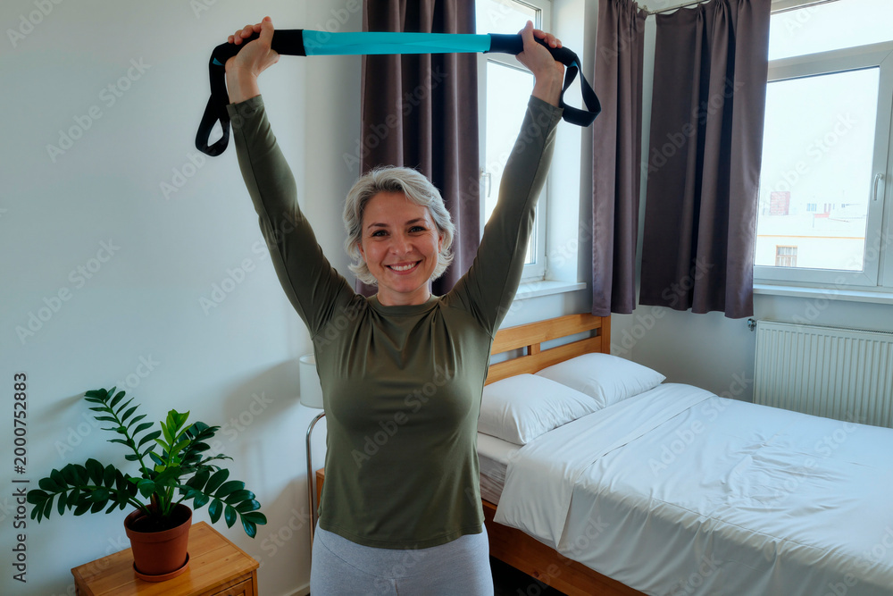 Fototapeta premium Portrait of adult woman stretching resistance band overhead at home, smiling and exercising for fitness and healthy aging. Useful for wellness, rehab, home workout marketing