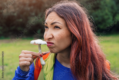Woman smelling a wild mushroom, showing a disgusted facial expression outdoors in nature, unsure about its edibility