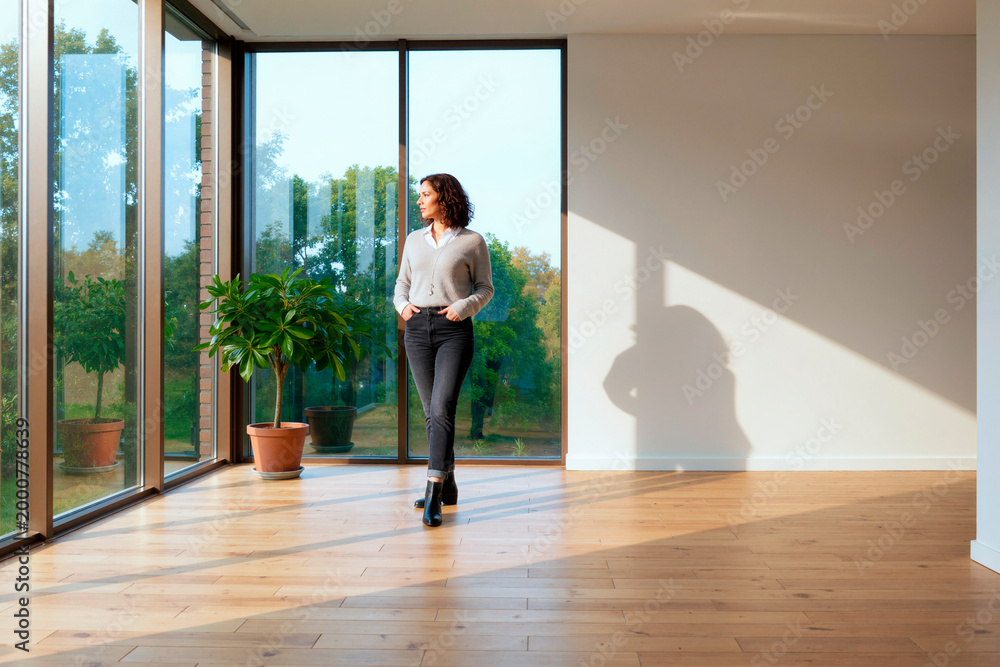 Fototapeta premium Wide shot of young woman standing near floor to ceiling windows in empty home, considering property move or renovation. Ideal for real estate, relocation, interior marketing