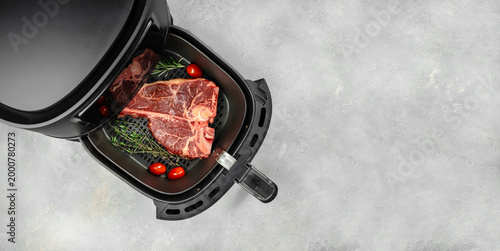 Konstfotografi Raw porterhouse steak in open air fryer basket with herbs and cherry tomatoes on light concrete background, top view