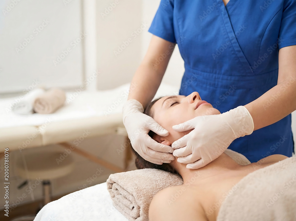 Obraz premium Professional in scrubs massaging head and neck of patient on treatment bed, clinical photo.