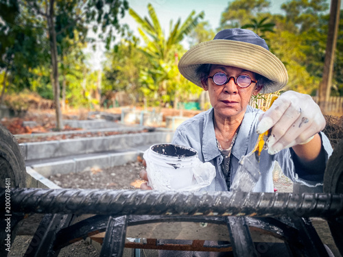 Senior woman painting metal at home garden, DIY repair and maintenance concept, wearing gloves and hat, focused lifestyle activity with copy space for text or image, natural outdoor background.