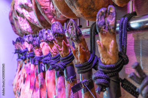 Side perspective of traditional Spanish Jamon Iberico legs hanging in a row. Close-up of Pata Negra hooves with black ropes in a gourmet cellar or deli with moody purple lighting