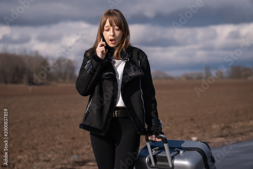 Foto Woman traveler pulling luggage bag down country road, communicating on smartphon
