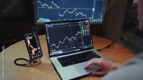 Man trader monitors market charts on laptop, screen and smartphone at home. Analyzing real-time trading data, considering investment strategies. Professional investor engaged in day trading, Evening