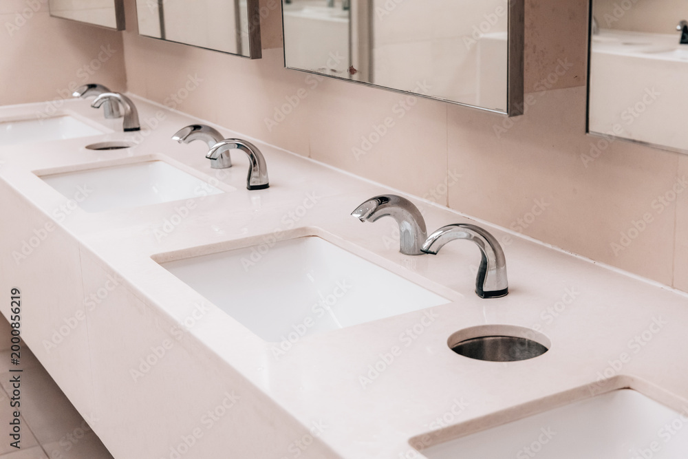 Fototapeta premium Multiple clean white sinks and automatic chrome faucets in a contemporary public bathroom, emphasizing hygiene and accessibility