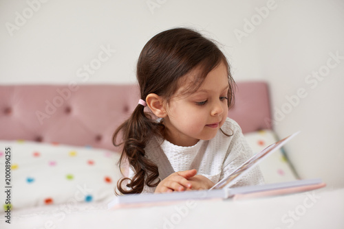 Little girl lying in bed, engrossed in reading a story book, showing early childhood education and concentration