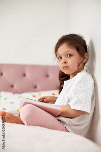 adorable child girl sitting in bed, holding a book and looking at the camera, promoting literacy and childhood education