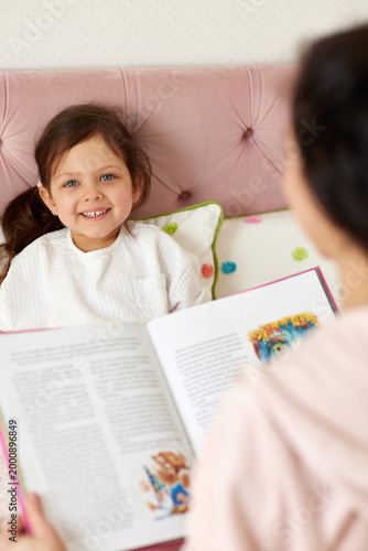 Young daughter smiling while her mother reads a book, sharing a moment of connection during a bedtime story
