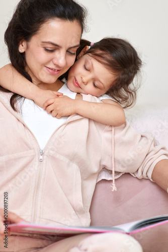 Young mother and happy daughter embracing closely while reading an interesting book together at home, sharing sweet moments