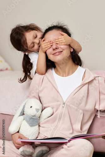 toddler daughter covering her mother's eyes from behind, playing a game while sitting on the floor with a rabbit toy and book