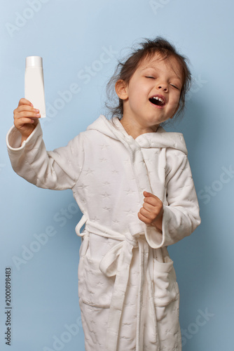 child girl in a white bathrobe holding a white lotion or cream tube, laughing with closed eyes