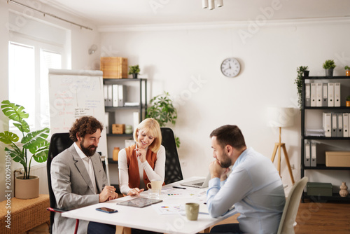 Three team members discuss ideas and strategies at a meeting table in a modern office while taking notes and sharing thoughts. Focus is on teamwork and collaboration.