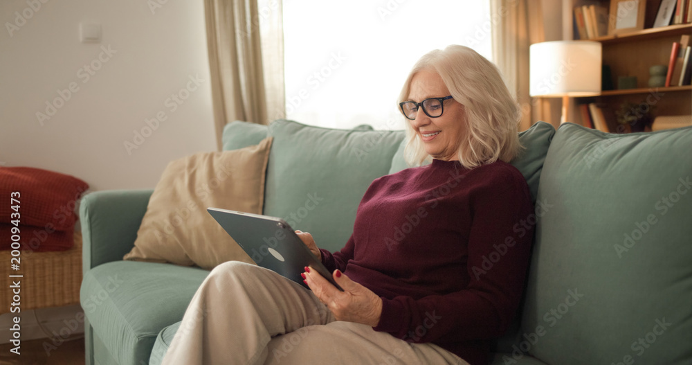 Obraz premium An older woman enjoys her time sitting on a couch while using a tablet. She wears glasses and has a smile. The room is warm with soft lighting and casual furniture.