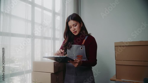 Asian small business owner checking inventory and writing on a clipboard in her home office with cardboard boxes ready for delivery