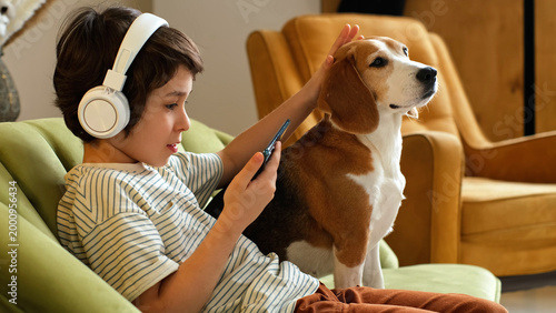 A boy listens through white headphones and looks at his phone while touching a beagle sitting close on a sofa. Digital habits, family pets, and quiet home routines create comfort and connection.