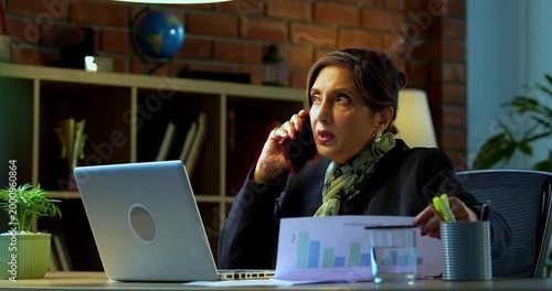 Senior Indian businesswoman in corporate attire attending phone call on smartphone while sitting with laptop and looking at documents in cozy office space experienced female executive at desk