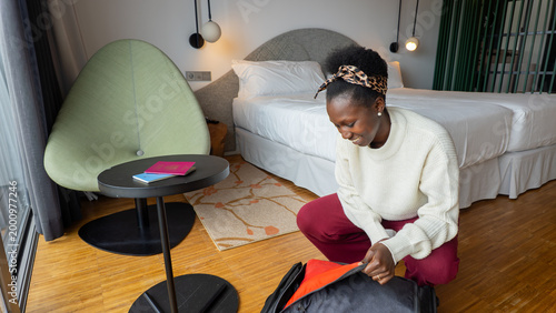 Tableau sur toile African traveler in a white sweatshirt and burgundy pants unpacking her gray suitcase in a hotel