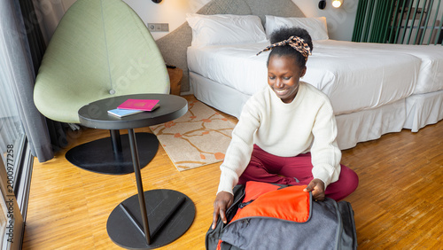 Tableau sur toile Young African woman in a white sweatsuit opening a gray suitcase in a modern hotel room